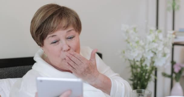 Feliz señora madura saludando comprar y soplar beso mientras termina la videollamada en la tableta. Vieja en casa robar tener video conversación durante el desayuno mientras está sentado en la cama con Trey . — Vídeo de stock