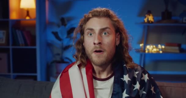 Close up view of handsome young man sitting in front of tv and supporting favourite team. Long haired guy with USA flaf on his shoulders watching sport and rejoicing victory. — Stock Video