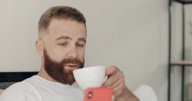 Genç sakallı adamın akıllı telefonunu kullanmasına ve sabahları kahve içerken gülümsemesine yakından bakın. Yakışıklı adam kahvaltı yaparken ve yatakta otururken telefon ekranına bakıyor..