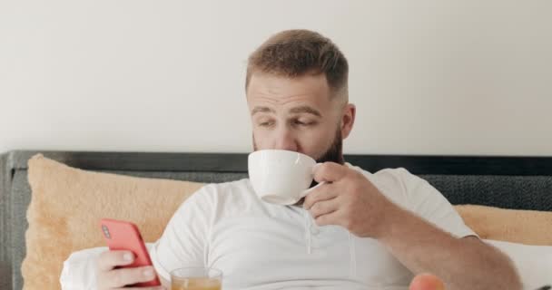 Vue rapprochée du bel homme barbu buvant du café au lit et souriant à la camée. Portrait de gars heureux dans les années 30 en utilisant son smartphone moderne pendant le petit déjeuner. Concept de technologie et de vie . — Video