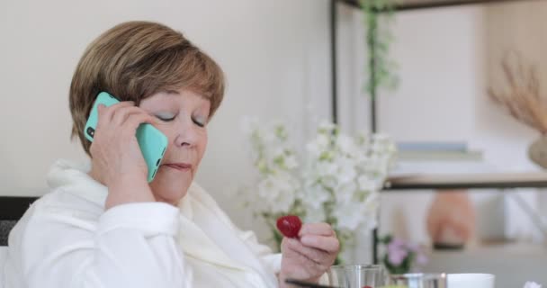 Close-up vista de boa aparência velha mulher falando e comendo morango enquanto toma café da manhã na cama. Senhora idosa em casa assaltar sentado e se comunicando no smartphone . — Vídeo de Stock