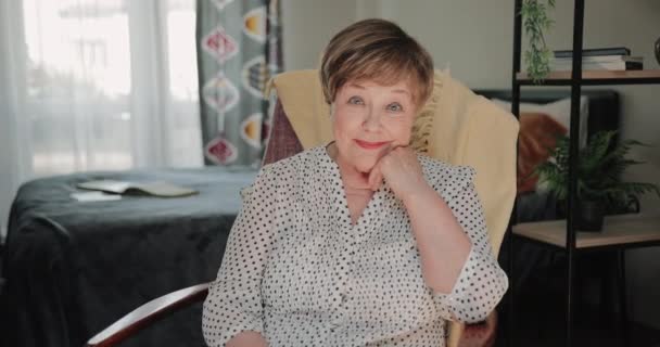 Retrato de una anciana guapa sentada en una silla y apoyando la cabeza en casa. Hermosa mujer jubilada sonriendo mientras mira a la cámara. Acércate. Concepto de personas y estilo de vida . — Vídeo de stock