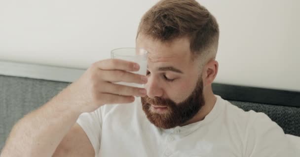 Homem barbudo nos anos 30 sofrendo de ressaca ang segurando vidro de água fria perto da cabeça. O jovem acordou de manhã e sentiu-se mal depois de beber a noite toda. Fundo de casa . — Vídeo de Stock