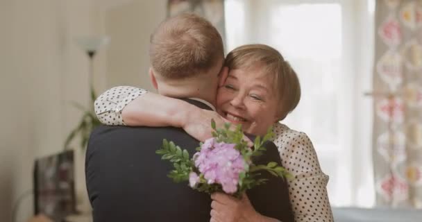Ευτυχισμένη όμορφη ώριμη μητέρα αγκαλιάζει τον ενήλικο γιο της κρατώντας λουλούδια στο χέρι της. Επισκέπτεται τη συνταξιούχο μητέρα του στο σπίτι. Έννοια της οικογένειας, της μητέρας ημέρα και τα γενέθλια. — Αρχείο Βίντεο