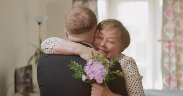 Feliz madre madura de buen aspecto abrazando a su hijo adulto mientras sostiene flores en su mano. Hombre visitando a su madre jubilada en casa. Concepto de familia, día de la madre y cumpleaños . — Vídeos de Stock