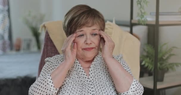 Äldre pensionär med huvudvärk och massagetempel. Kvinna i 70-talet i ser trött och gnugga tempel när du sitter i stol hemma. Begreppet människor och hälsoproblem. — Stockvideo
