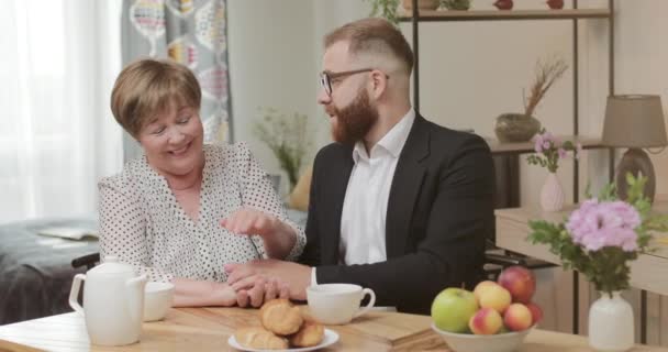 Glad mogen mamma och vuxen son pratar och kopplar av medan de sitter i köket tillsammans.Ung affärsman besöker sin vackra mamma. Begreppet kärlek och omsorg. — Stockvideo