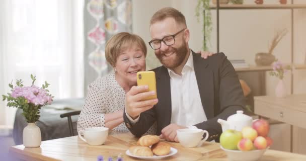 Radosny dorosły syn i dojrzała matka wykonują wideo połączenie ze smartfonem siedząc w domu. Starsza kobieta i młody mężczyzna za pomocą aparatu czołowego nowoczesnego nobile telefon, uśmiechając się i machając. — Wideo stockowe