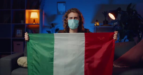 Odpowiedzialny młody człowiek w masce ochronnej trzymający włoską flagę siedząc na sofie. Tysiąclecia facet zostaje w domu z powodu kwarantanny.Koncepcja wybuchu epidemii patogenu koronawirusa. — Wideo stockowe