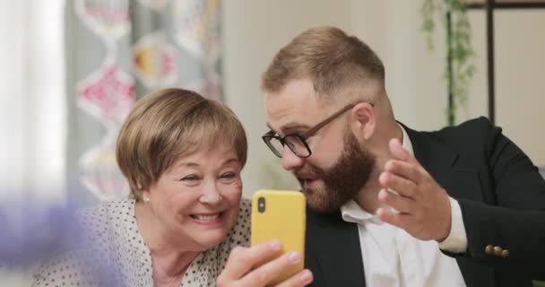 Oudere vrouw en volwassen kleinzoon die een videogesprek hebben met familie. Jongeman die zijn oma videocall leert maken terwijl hij haar thuis bezoekt. Concept van technologie en communicatie. — Stockvideo