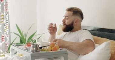 30 'lu yaşların ortasında, yatakta kahvaltı yaparken ilaç alan sakallı bir adam. Genç adam tepsi dolusu yiyecekle otururken bir bardak suyla hap içiyor. Tıp ve sağlık hizmetleri kavramı.