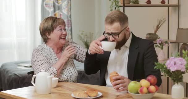 Snygg man i glas och kostym som äter och dricker när han sitter vid mödrarnas bord hemma. Glada pensionerad kvinna på 70-talet ser glad och skrattar medan du spenderar tid med son. — Stockvideo