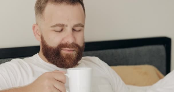 Blisko widok przystojnego białego mężczyzny po trzydziestce, pachnącego kawą. Portret brodatego faceta po trzydziestce, cieszącego się porankiem leżącego w łóżku i uśmiechniętego do kamery. Kontekst domu. — Wideo stockowe