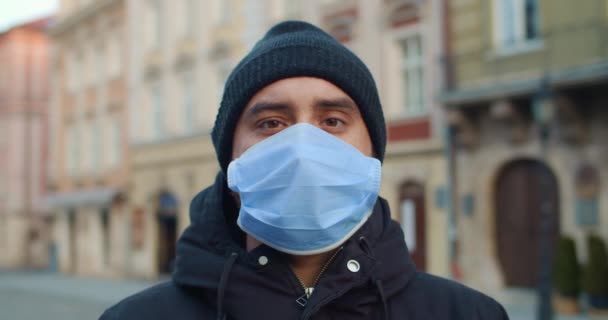 거리에 서 있는 카메라를 찾기 위해 보호용 마스크를 쓴 남자의 모습을 가까이 서 볼 수있다. 얼굴에 의료 마스크를 쓴 젊은 남자의 초상화. 텅 빈 유럽의 도시 가 배경에 있습니다. 유행병에 대한 개념. — 비디오