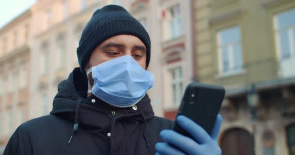 Visão de perto do homem com máscara protetora e luvas olhando para a tela do telefone. Cara com máscara médica um em seu rosto de pé sozinho e segurando smartphone. Conceito de pandemia de coronavírus . — Vídeo de Stock