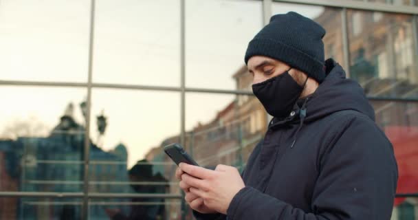 Visão lateral do cara digitando mensagem enquanto está na rua.Homem com máscara protetora de algodão preto no rosto usando smartphone enquanto se comunica nas redes sociais. Conceito de pandemia . — Vídeo de Stock