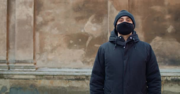 Портрет молодого человека в черной хлопчатобумажной защитной маске, стоящего перед старой стеной здания. Концепция жизни и безопасности, коронавирус, защита от вирусов, мир пандемии . — стоковое видео