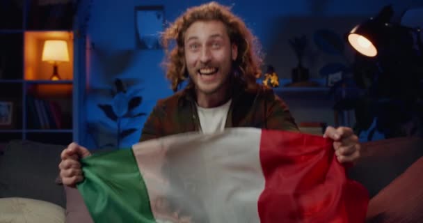 Milli takımının başarısını kutlayan heyecanlı genç adam ve çok mutlu görünüyor. Yakışıklı bir adam oturma odasında televizyon izlerken elinde İtalyan bayrağı ve sevinç dolu bir bayrak tutuyor.. — Stok video