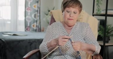 Evinde örgü iğneleriyle sandalyede oturan güzel büyükannenin ön görüntüsü. Neşeli yaşlı bayan boş zaman harcıyor ve örgü örüyor. Boş zaman ve emeklilik kavramı.