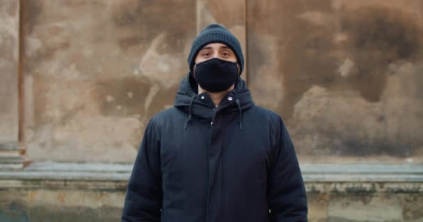 Πορτρέτο του νεαρού άντρα με τη μαύρη προστατευτική βαμβακερή μάσκα στέκεται μπροστά από το παλιό κτίριο τοίχο. Έννοια της ζωής για την υγεία και την ασφάλεια, coronavirus, προστασία από ιούς, πανδημία στον κόσμο. — Αρχείο Βίντεο