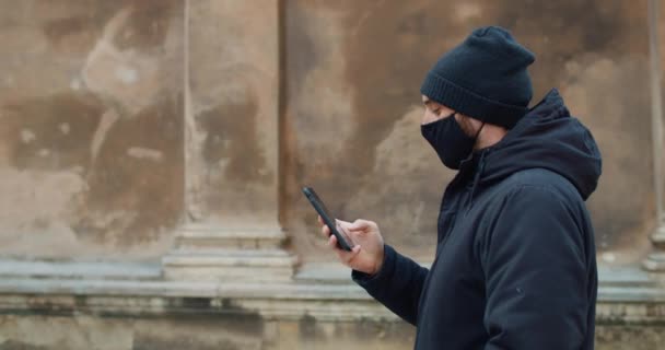 Vista lateral do homem andando e usando seu smartphone para se comunicar em redes sociais. Cara de algodão preto máscara protetora digitando mensagem e olhando para a tela do telefone. Conceito de coronovírus . — Vídeo de Stock