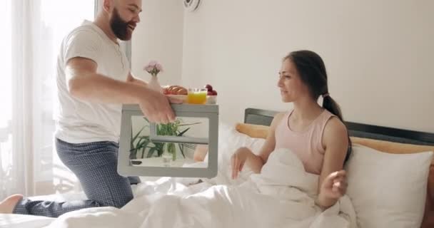 30代のハンサムな男が妻のためにベッドで朝食を取り、彼女の近くに座っています。幸せなガールフレンドは彼女のボーイフレンドが食べ物でトレイを保持し、彼にキスを待っている。ライフスタイルの概念. — ストック動画