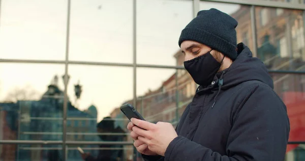 Side view of guy typing message while standing at street.Man with black cotton protective mask on face using smartphone while communicating in social networks. Concept of pandemic. — Stock Photo, Image