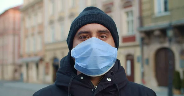 Close up view of man with protective mask looking to camera standing at street. Portrait of young guy in medical mask on his face. Empty old europian city at background. Concept of pandemic. Stock Image