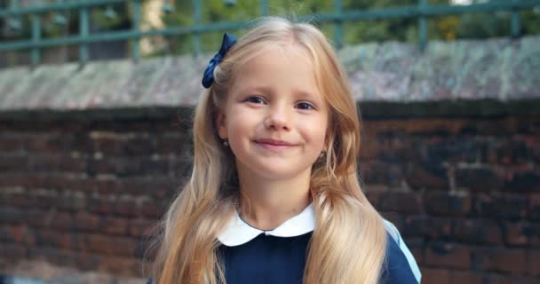 Ritratto di bambina in uniforme blu che guarda alla telecamera. Vista da vicino di bella studentessa con lunghi capelli biondi e zaino sorridente mentre si trova in strada . — Video Stock