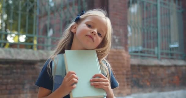 Portrait de mignonne petite écolière tenant livre et regardant à la caméra. Gros plan d'adorable enfant aux longs cheveux blonds frappant et posant tout en se tenant debout dans la rue. Concept d'enfant . — Video