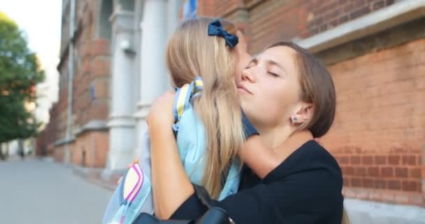 Närbild av liten bedårande flicka omfamna sin mamma innan du springer till skolan byggnad. Ung kvinna pratar och vinkar till sin dotter som går på lektioner på morgonen. — Stockvideo