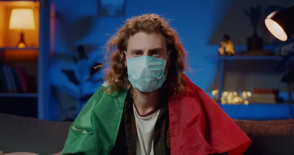 Millenial man in protective mask on face with italian flag on shoulders looking to camera. Portrait of guy staying at home on quarantine. Concept of safety life, coronavirus, virus protection. Stock Picture