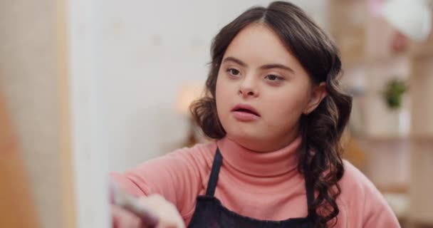 Vista da vicino della ragazza talentuosa con la sindrome di Down nella pittura quadro e sorridente.Bella adolescente guardando felice mentre seduto di fronte a molbert. Concetto di bambini soleggiati. Interni . — Video Stock