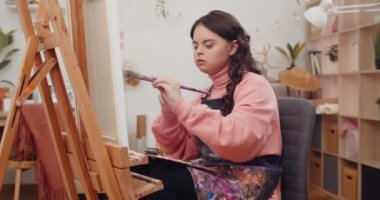 Ressam önlüğü giyen ve fırçayı bir elinden diğer eline koyan bir genç. Molbert 'in önünde resim yaratırken dizlerinin üzerinde palet tutan Down sendromlu bir kız..