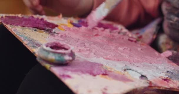 Bekijk van dichtbij het kunstenaarspalet met roze, gele, witte en paarse kleuren erop. Kunstpalet met gemengde vlekken en penseel weken in verf. Concept van creativiteit en schilderkunst. — Stockvideo