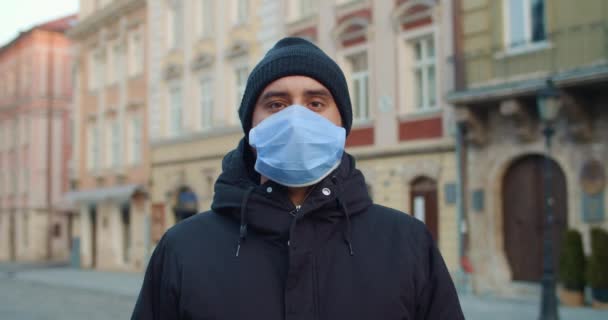 Crop view of man in protective mask standing at empty street. Portrait d'un gars avec un masque médical sur le visage regardant vers une caméra dans une vieille ville européenne. Concept de pandémie de coronavirus . — Video
