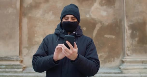 Vooraanzicht van de mens die loopt en zijn smartphone gebruikt voor communicatie in sociale netwerken. Man in zwart katoen beschermend masker typt bericht en kijkt naar het telefoonscherm. Concept van COVID 19. — Stockvideo