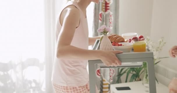 Hermosa mujer que lleva el desayuno en la cama para el marido, besándose y sentado cerca de él. Feliz novio esperando a su novia sosteniendo bandeja con comida. Concepto de estilo de vida y relación . — Vídeo de stock