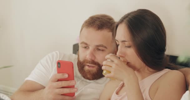 Vista de perto do casal feliz falando enquanto olha para a tela do telefone. Mulher bebendo suco enquanto o homem segurando smartphone durante o café da manhã na cama. Conceito de relacionamento e lazer — Vídeo de Stock