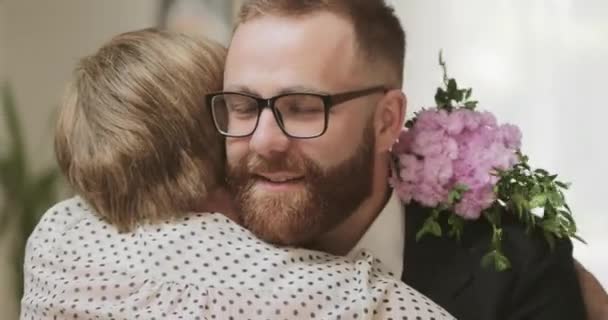 Vista de cerca de un hombre adulto sonriente con gafas abrazando a su abuela y trayéndole flores. Joven guapo visitando a su madre pensionista en casa.Concepto de familia, día de las madres . — Vídeo de stock