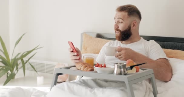 Joven sorprendido usando su teléfono inteligente y tener buenas noticias. Hombre adulto hombre de negocios de 30 años desayunando y tomando café mientras recibe noticias impactantes. Concepto de tecnología y vida . — Vídeos de Stock