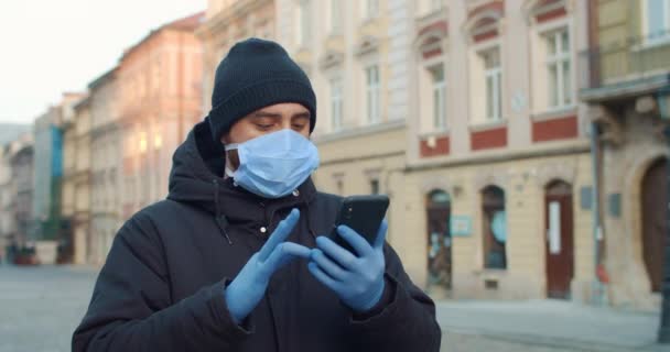 Człowiek przewijania wiadomości i patrząc na ekranie telefonu podczas spaceru po pustej ulicy. Facet w medycznej masce ochronnej i rękawiczkach używający smartfona w starym europejskim mieście. Pojęcie koronawiru. — Wideo stockowe