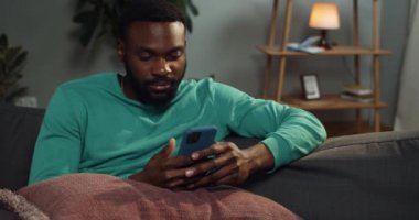 Evde vakit geçirirken akıllı telefon kullanan yakışıklı sakallı adamın portresi. Genç Afro-Amerikalı adam evdeki kanepede otururken telefon ekranına bakıyor ve internette geziniyor..