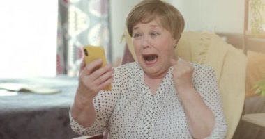 Olgun, neşeli bir bayan harika haberler veriyor ve son derece mutlu görünüyor. Yaşlı kadın emekliler modern akıllı telefonunu kullanıyor ve sevinçle zafere koşuyorlar. Başarı ve teknoloji kavramı.