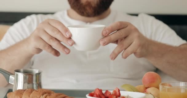 Szczęśliwy młodzieniec wąchający kawę w łóżku i uśmiechający się. Wesoły brodaty facet jedzący śniadanie siedząc z tacą pełną jedzenia. Pojęcie "dzień dobry" .Home background. — Wideo stockowe