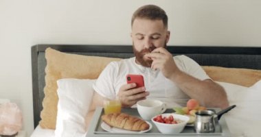 Ciddi sakallı adam akıllı telefonunu kullanıyor ve yatakta otururken elma yiyor. 30 'lu yaşlarda yakışıklı bir adam yemek dolu bir tepside oturup telefon ekranına bakarken kahvaltı yapıyor..