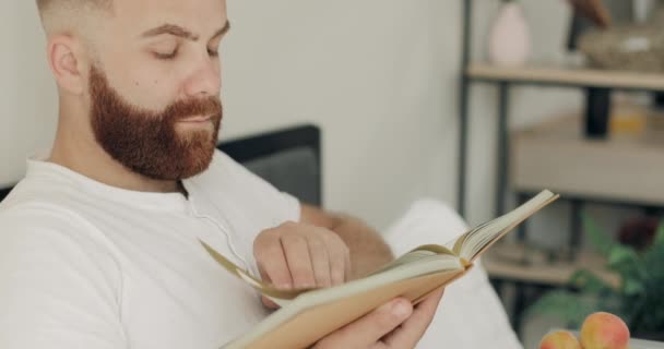 Close-up zicht op geconcentreerde jongeman die roman leest tijdens het ontbijt op bed. Knappe man met een baard in het midden van de 30, zittend en draaiend. Het begrip vrije tijd. — Stockvideo