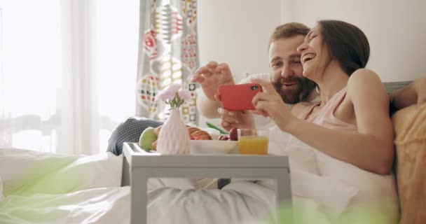 즐거운 가족들은 쟁반에 음식 이 가득 들어 있는 침대 위에 앉아 사진을 보고 있다. 젊은 남녀가 침대에서 아침을 먹으면서 스마트폰 화면을 보고 만지면서 웃고 있습니다. — 비디오