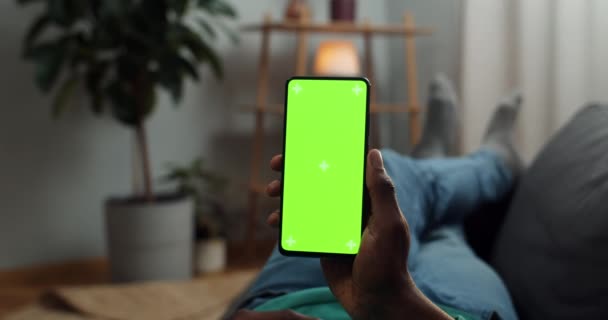 Hombre joven acostado en el sofá con un teléfono inteligente moderno en la mano mientras mira en la pantalla de la maqueta con marcadores de demolición en él. Concepto de pantalla verde y croma key. Acogedor fondo casero . — Vídeos de Stock