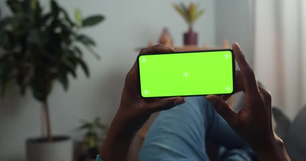 Afrykański facet przesuwając smartfon makieta ekran z markerów trecking trzymając go w poziomym trybie krajobrazu. Facet gra w grę leżąc na kanapie. Pojęcie klucza chromatycznego. — Wideo stockowe
